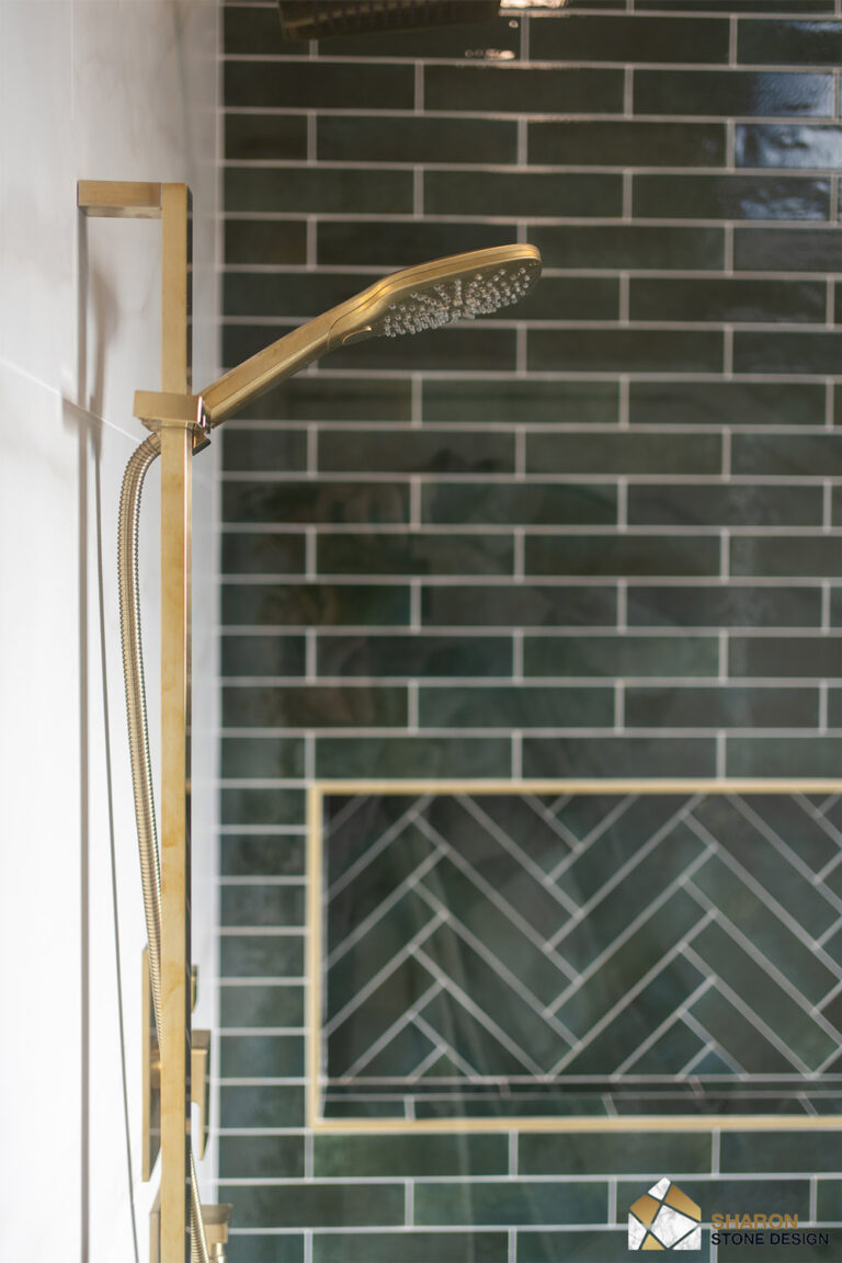Palo Alto bathroom remodel handheld shower in brushed brass mounted on a tall slide bar, photographed through the glass panel with the herringbone shampoo niche and green tile behind.