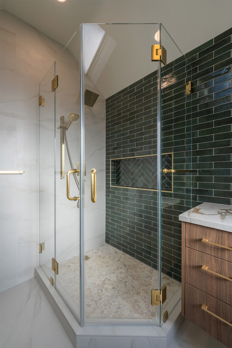 Palo Alto bathroom remodel corner shower with brass hinges and handles on frameless glass, a brushed brass rain head and handheld, deep green subway tile with a herringbone shampoo niche trimmed in brass Schluter profile, and a cream pebble mosaic floor.