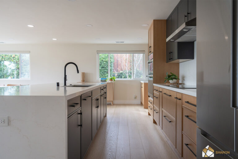 Redwood City home remodel kitchen looking down the white quartz island with its matte black pull-down faucet, charcoal island cabinetry, and a white oak run opposite holding an induction cooktop beneath a stainless hood.