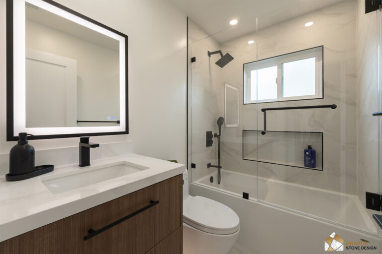 Redwood City home remodel guest bathroom with a black-framed backlit mirror over a walnut floating vanity, quartz top and black faucet, alongside a glass-screened tub with black fittings, a shampoo niche trimmed in black Schluter profile and a grab bar.