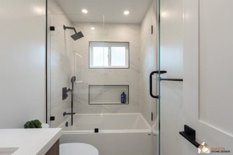 Redwood City home remodel tub alcove viewed head-on, with a fixed glass screen, black rain head and handheld, a window and shampoo niche framed in black Schluter profile against marble-look porcelain, and a black towel bar at right.