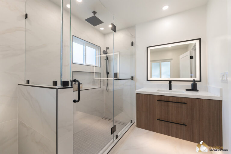Redwood City home remodel primary bathroom from the doorway, with the glass shower and half-wall at left and a walnut floating vanity with two black bar pulls beneath a black-framed lit mirror at right.