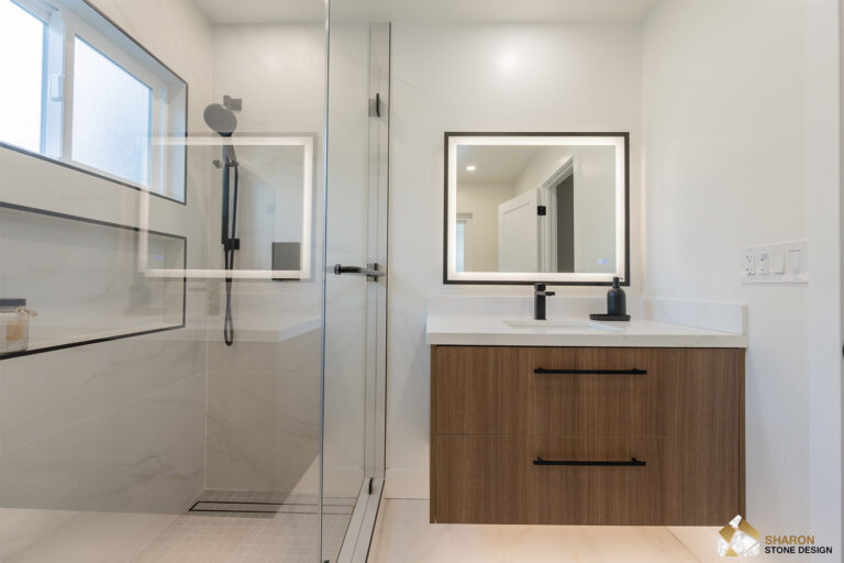 Redwood City home remodel bathroom looking across the shower glass to the walnut vanity, its quartz top and black faucet lit by a black-framed LED mirror, with the shampoo niche and handheld reflected in the panel.