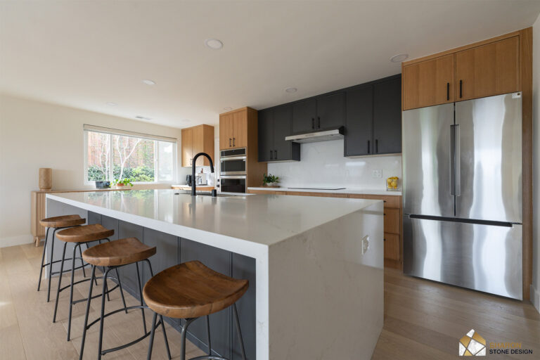 Redwood City home remodel kitchen from the dining end, the long quartz waterfall island leading toward charcoal and white oak cabinetry, a stainless chimney hood, and a stainless refrigerator in an oak surround.
