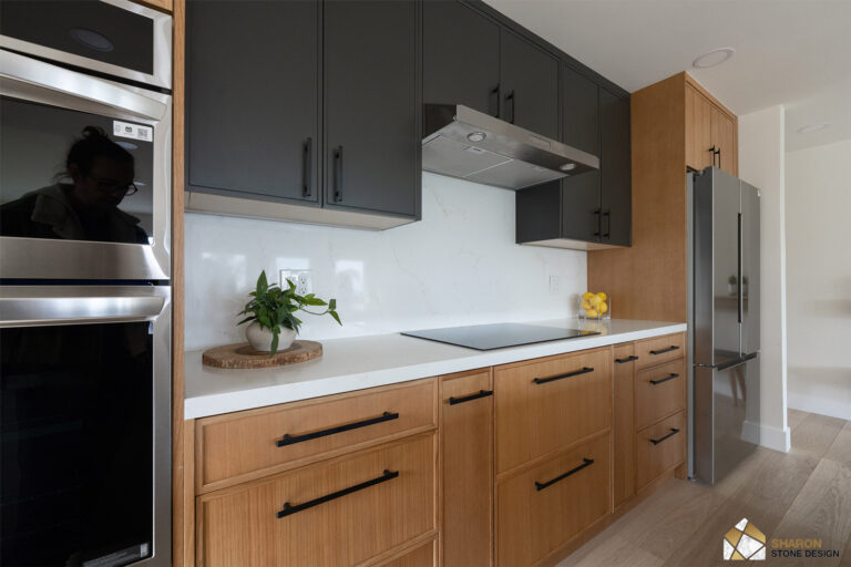 Redwood City home remodel cooking run with a flush induction cooktop set in quartz, a stainless under-cabinet hood, a quartz slab backsplash, and charcoal uppers above white oak drawers with long black pulls.
