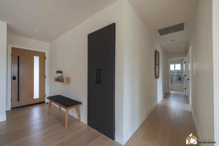 Redwood City home remodel entry hall with a white oak front door and long black pull, a floating oak shelf, an oak bench with a charcoal cushion, and full-height charcoal closet doors beside the corridor to the bathroom.
