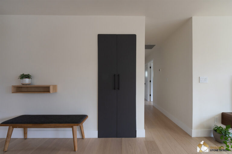 Redwood City home remodel entry storage closed behind flush charcoal doors with slim black pulls, flanked by a floating oak shelf and cushioned bench on pale oak flooring.
