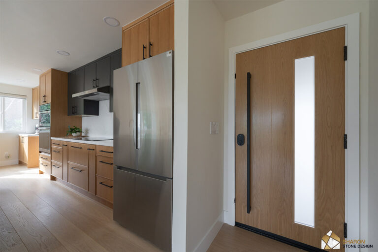 Redwood City home remodel looking from the front door toward the kitchen, past the stainless refrigerator in its oak surround, with the white oak entry door and its frosted vision panel at right.