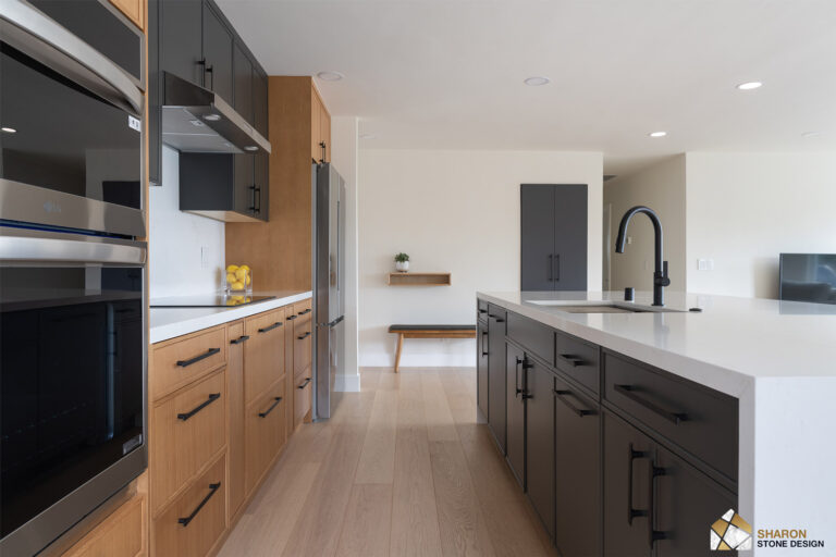 Redwood City home remodel kitchen seen past stainless double wall ovens, with charcoal upper cabinets above white oak drawers, a stainless refrigerator, and the quartz island leading to an oak bench in the hall beyond.