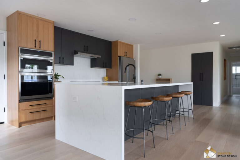 Redwood City home remodel island with a mitered quartz waterfall end, charcoal cabinetry, and a white oak tower housing stainless double wall ovens.