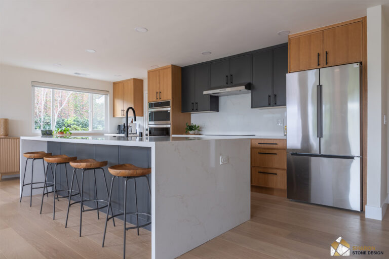 Redwood City home remodel kitchen from the living room, showing the full length of the quartz island, the two-tone oak and charcoal cabinetry, a stainless refrigerator, and a fluted oak sideboard beneath the window.