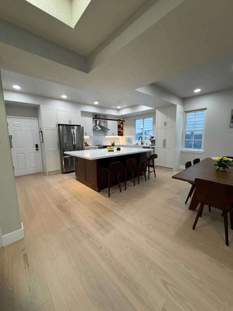 Redwood City home remodel open plan seen from the entry, with the dark walnut veneer island at center, and white shaker cabinetry and stainless appliances beyond.