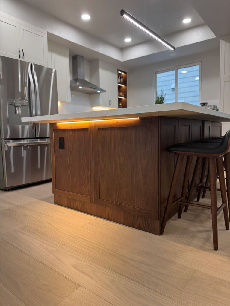 Redwood City home remodel island end in walnut paneling with a concealed LED strip washing the floor beneath the quartz overhang, alongside stainless refrigeration and a black linear pendant.