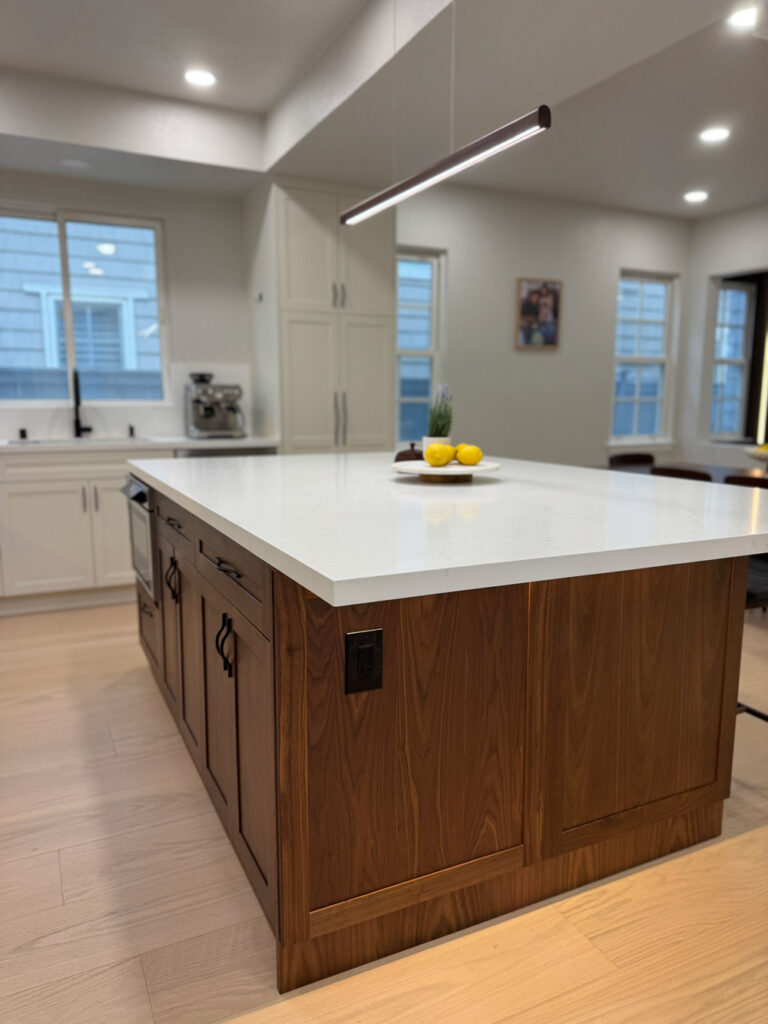 Redwood City home remodel island corner from a low angle, the white quartz top mitered over walnut cabinetry fitted with black pulls and an integrated power outlet, with the dining room windows beyond.