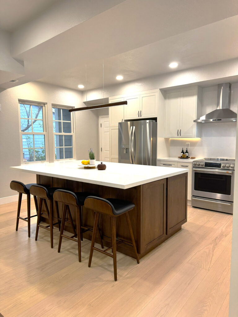 Redwood City home remodel kitchen from the living room side, the walnut island set against white shaker cabinetry, a stainless slide-in range and chimney hood, and daylight from the double windows.