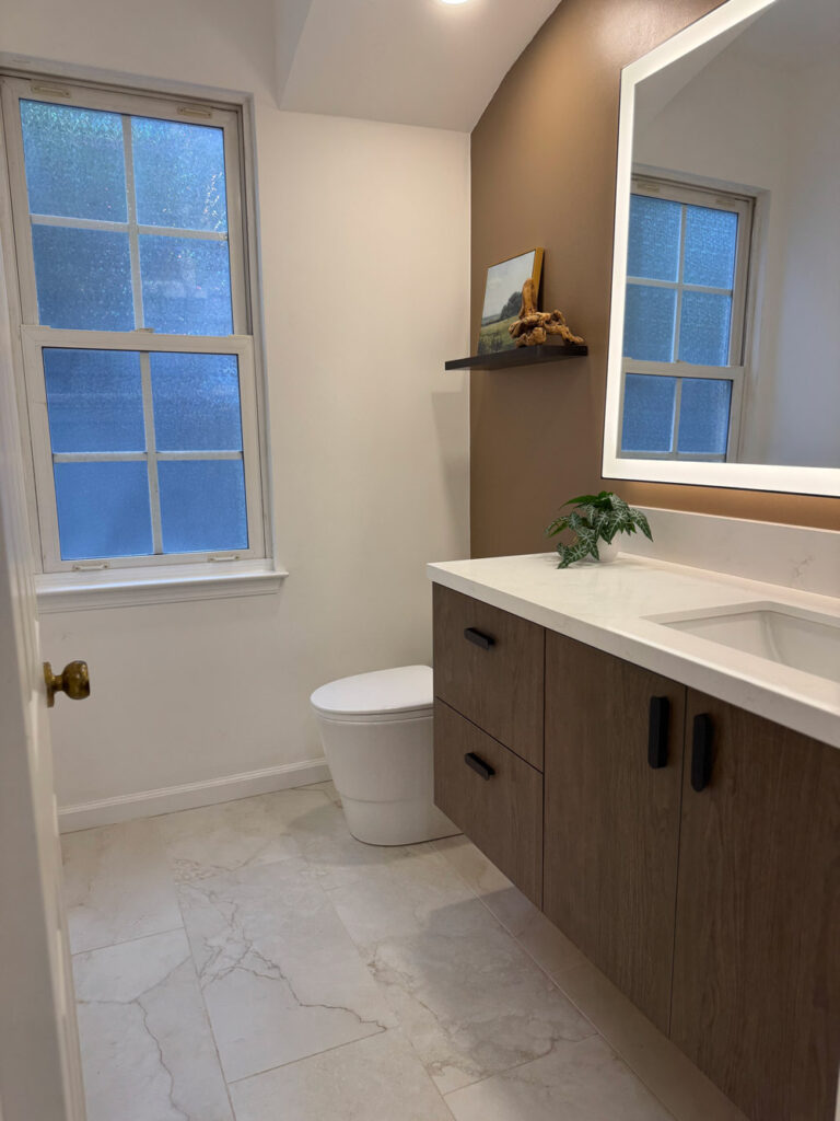 Redwood City home remodel guest bathroom with an obscured-glass sash window, a dark oak floating vanity with black bar pulls and white quartz top, a backlit mirror, and a mocha accent wall carrying a black floating shelf.