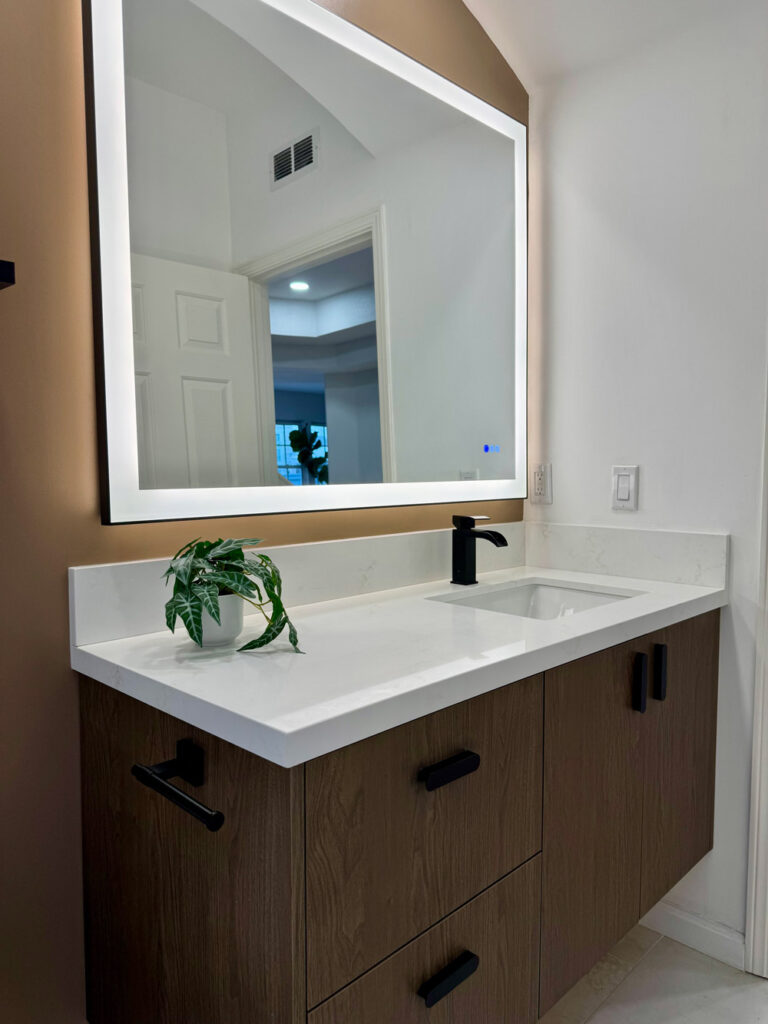 Redwood City home remodel bathroom vanity in dark oak with black bar pulls, a white quartz top and integrated upstand, a matte black faucet, and a large backlit mirror reflecting the hallway.