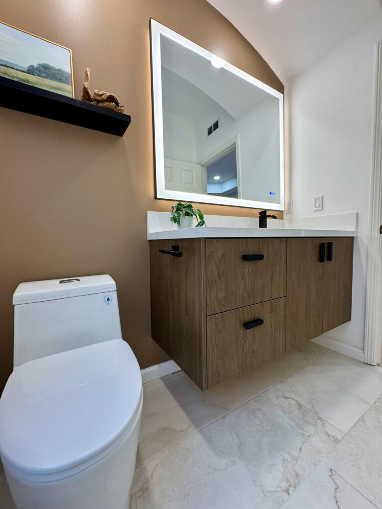 Redwood City home remodel bathroom seen past the toilet, with the black floating shelf on the mocha wall, the lit mirror above the floating oak vanity, and marble-look floor tile with soft veins throughout.