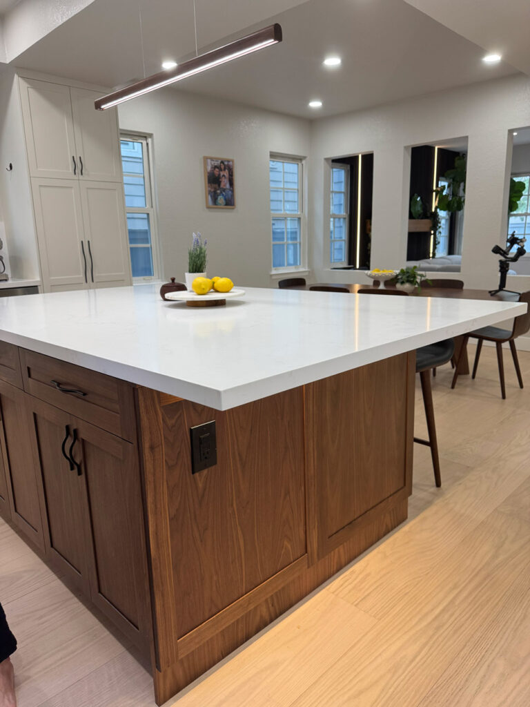 Redwood City home remodel island viewed from the dining side, its walnut paneled base fitted with black pulls and a power outlet, with the glowing slat wall of the living room beyond.