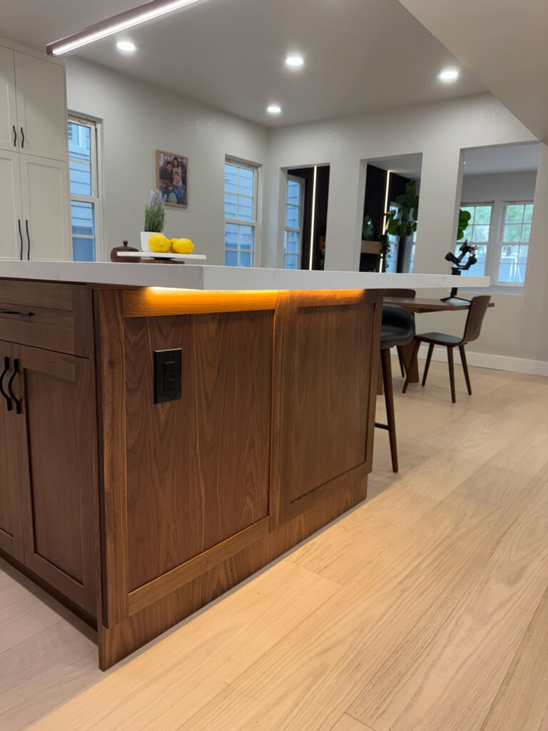 Redwood City home remodel island end lit by a warm LED reveal beneath the quartz overhang, its walnut paneling carrying a black power outlet, with interior windows to the living room beyond.