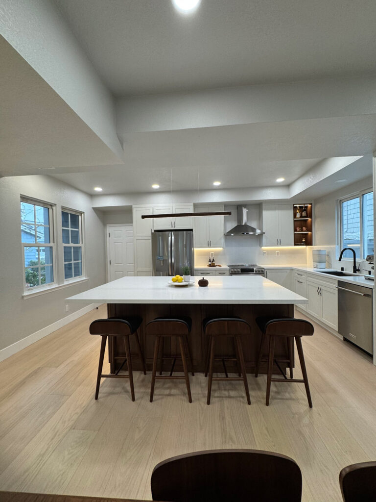 Redwood City home remodel kitchen viewed symmetrically down the room, the quartz island centered between white shaker cabinetry, a stainless refrigerator, chimney hood and range.