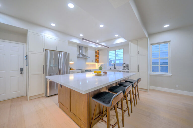 Redwood City home remodel kitchen with white shaker cabinetry, a generous white quartz island on a walnut base, a stainless refrigerator and chimney hood, a slim linear pendant, and an LED-lit walnut display niche.