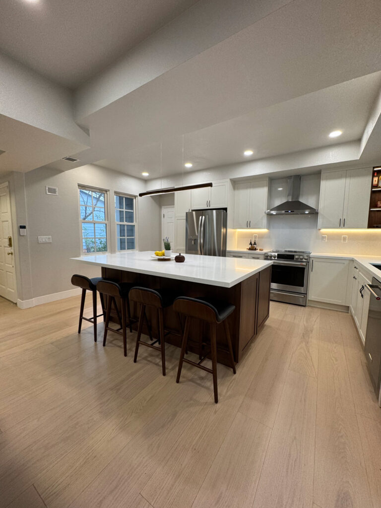 Redwood City home remodel kitchen from the dining corner, the walnut island angled across pale oak flooring beneath a linear pendant, with stainless appliances lining the far wall.