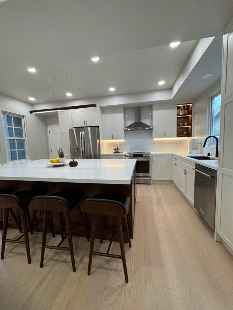 Redwood City home remodel kitchen with the island at left and the sink run with its black faucet and stainless dishwasher at right, framed by the range, chimney hood and LED-lit walnut display niche.
