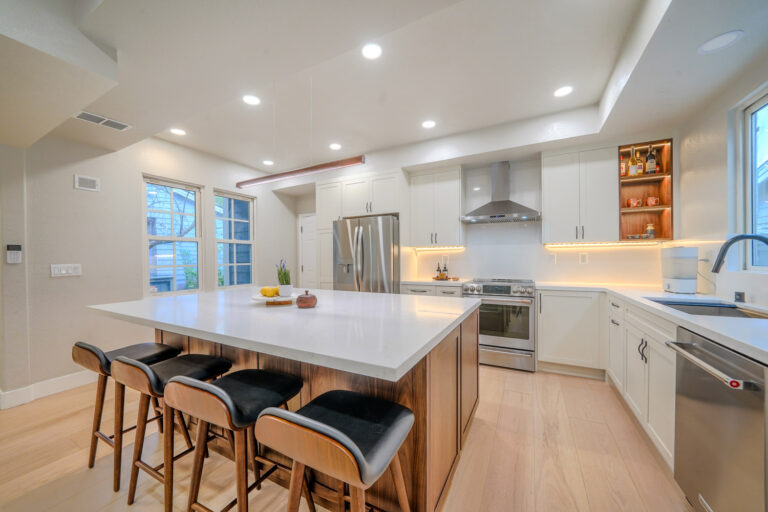 Redwood City home remodel kitchen viewed from the dining end, the quartz island leading to the stainless range, chimney hood and LED-lit walnut display niche beyond.