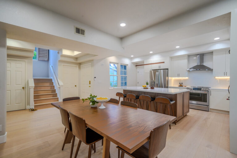 Redwood City home remodel open plan with the quartz island and white shaker kitchen at center, and the staircase and entry door at left on pale oak flooring.