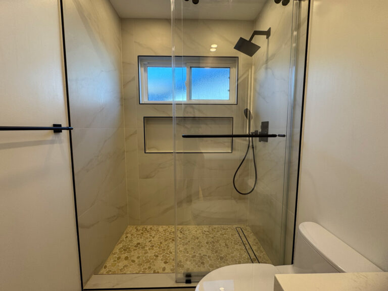 San Jose bathroom remodel shower viewed square through the glass, with an obscured window glowing blue above a shampoo niche trimmed in black Schluter profile, a matte black rain head and handheld, and a warm pebble mosaic floor.