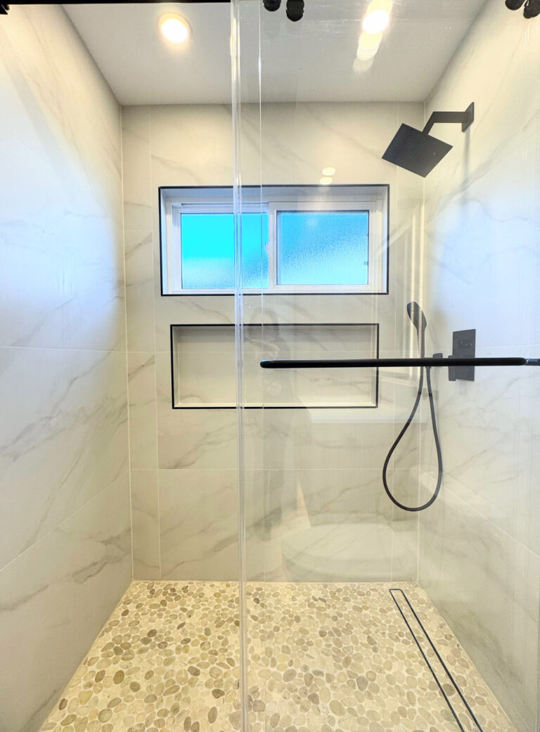 San Jose bathroom remodel shower enclosure in portrait view, the porcelain walls with soft veins framing a shampoo niche and obscured window trimmed in black Schluter profile, with a coiled black handheld hose and a linear drain in the pebble floor.