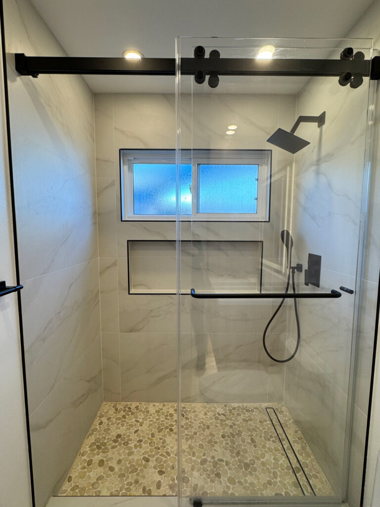 San Jose bathroom remodel shower with the full black barn-style roller rail spanning the top of the glass, a shampoo niche trimmed in black Schluter profile, matte black fittings, and a pebble floor draining to a linear channel.