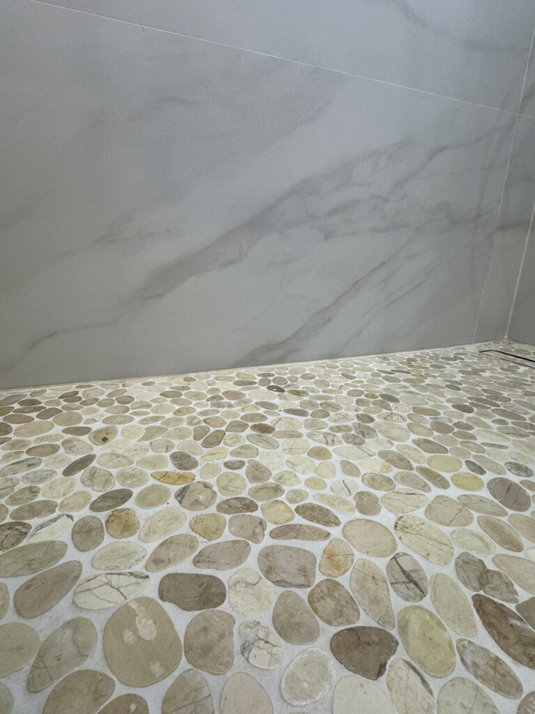 San Jose bathroom remodel shower floor detail where the tumbled pebble mosaic in cream and taupe meets the Calacatta-look wall panels with soft veins in the corner.
