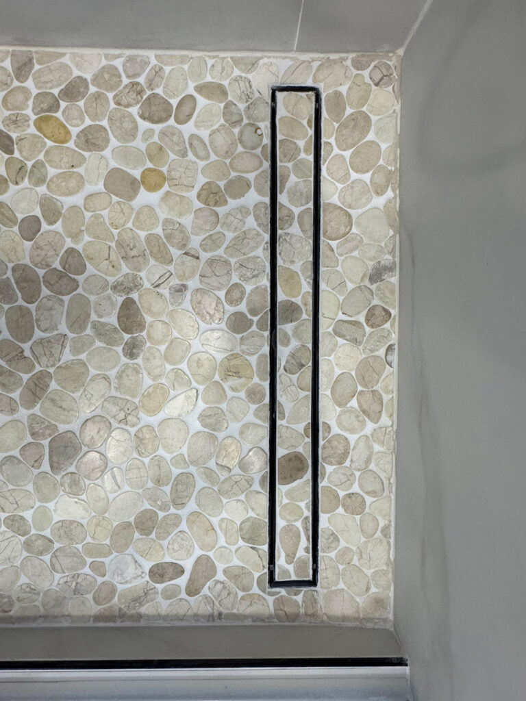 San Jose bathroom remodel overhead detail of the pebble mosaic shower base, its rounded stones running up to a slim black linear channel drain set flush with the floor.