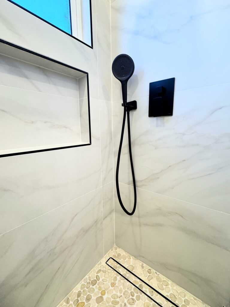 San Jose bathroom remodel shower corner with a round matte black handheld on its bracket, a black square pressure-balance valve, and the shampoo niche and window edges trimmed in black Schluter profile above the pebble floor.