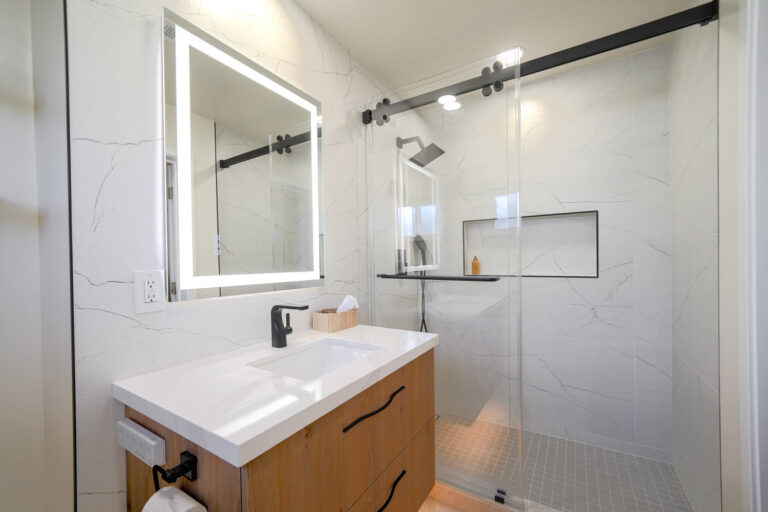 San Jose bathroom remodel with a backlit LED mirror above a walnut floating vanity, a matte black faucet, and a glass shower on a black barn-style rail lined in porcelain with soft veins and a shampoo niche trimmed in black Schluter profile.