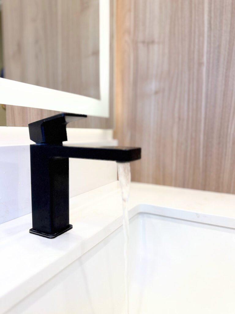 San Jose bathroom remodel faucet detail, a matte black square-spout mixer running water into the undermount basin, with the walnut vanity and lit mirror softly blurred behind.