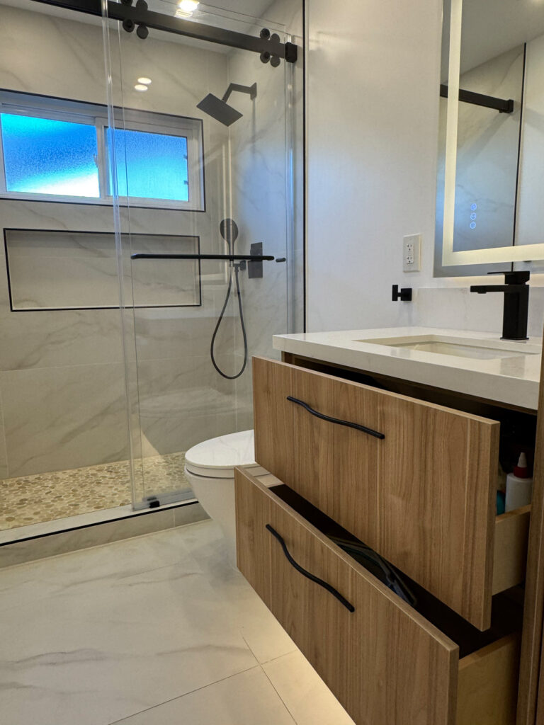San Jose bathroom remodel vanity with both walnut drawers pulled open to show the internal storage, their wavy black pulls catching the light, with the shower and toilet beyond.