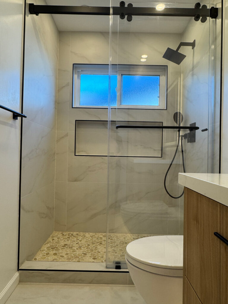 San Jose bathroom remodel shower on its black sliding rail with an obscured blue-lit window above the shampoo niche, matte black rain head and handheld, and the toilet and walnut vanity edge at right.
