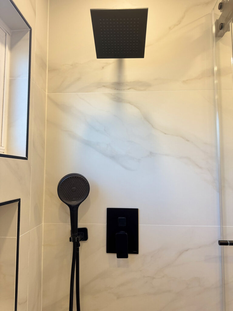 San Jose bathroom remodel shower wall detail with a large matte black square rain head, a round handheld on its bracket, and a black pressure-balance valve set against porcelain with soft veins.