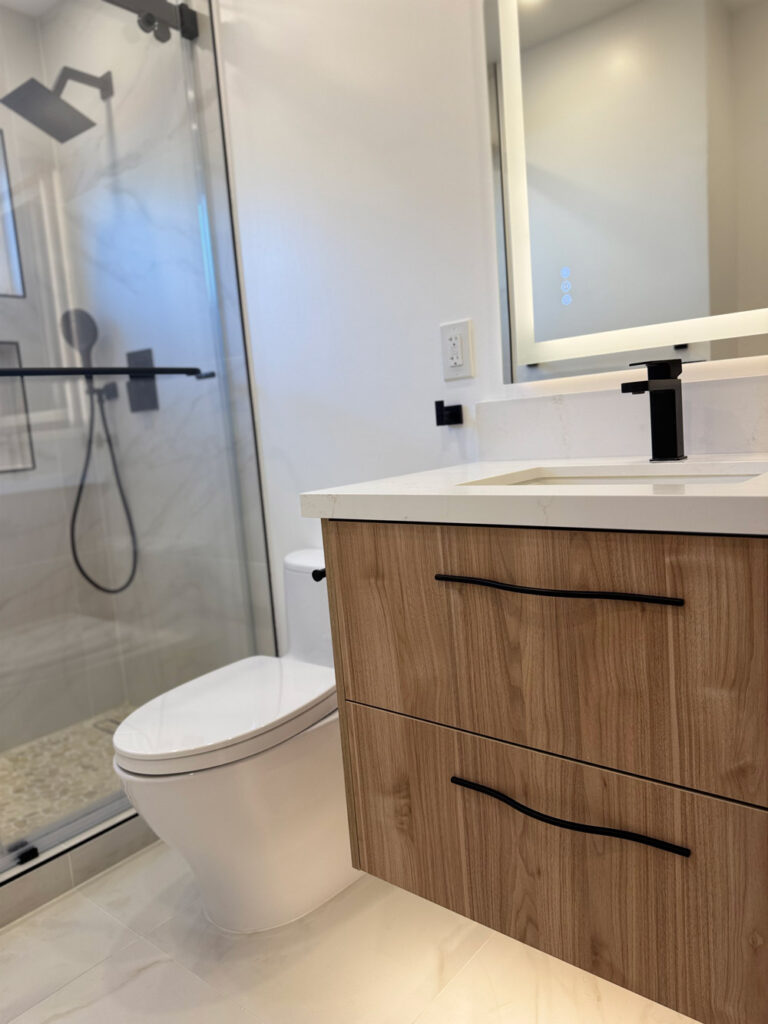 San Jose bathroom remodel looking along the walnut vanity toward the toilet and glass shower, the quartz counter and black faucet lit by the backlit mirror above.