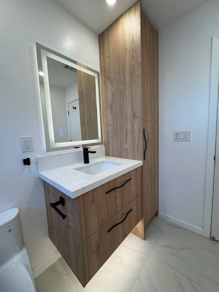 San Jose bathroom remodel master bath with a pale oak floating vanity, quartz top and black faucet below a backlit mirror, a black paper holder, and a full-height oak storage tower beside the toilet.