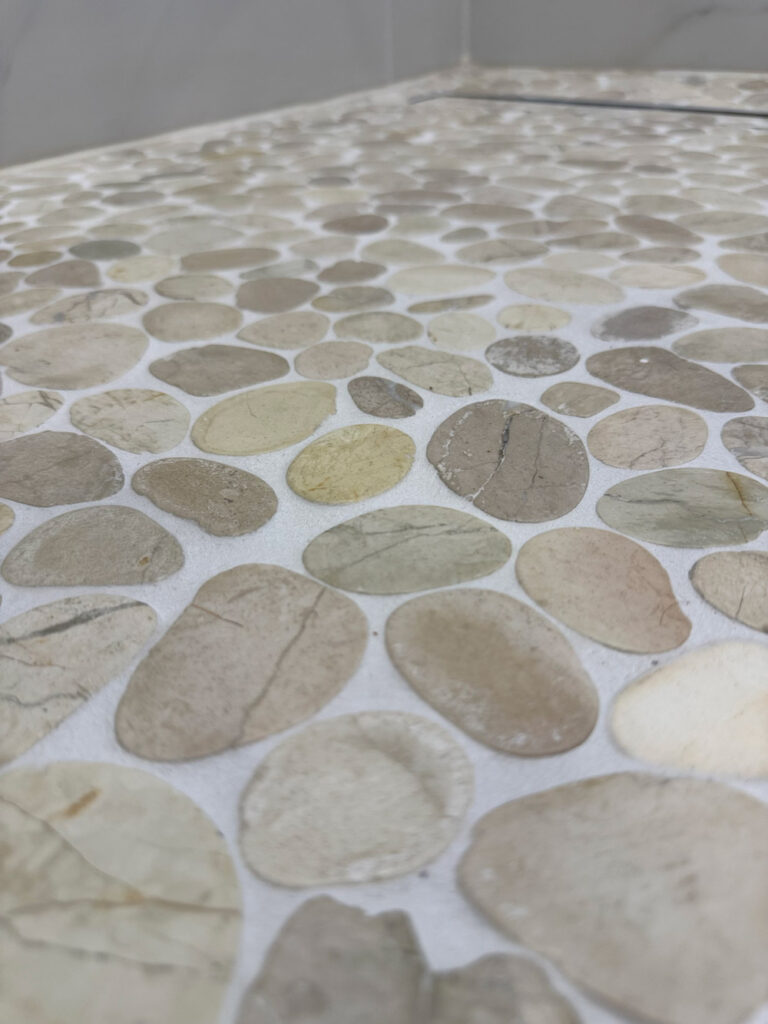 San Jose bathroom remodel angled close-up of the pebble shower floor, showing the range of cream, olive and gray tones and the fine veins running through individual stones.