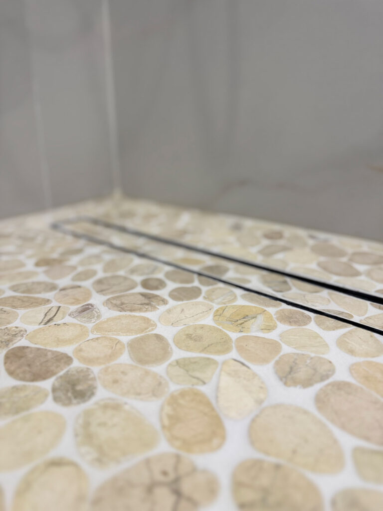 San Jose bathroom remodel pebble floor photographed along the black linear drain, the channel running diagonally between the rounded stones toward the porcelain wall.