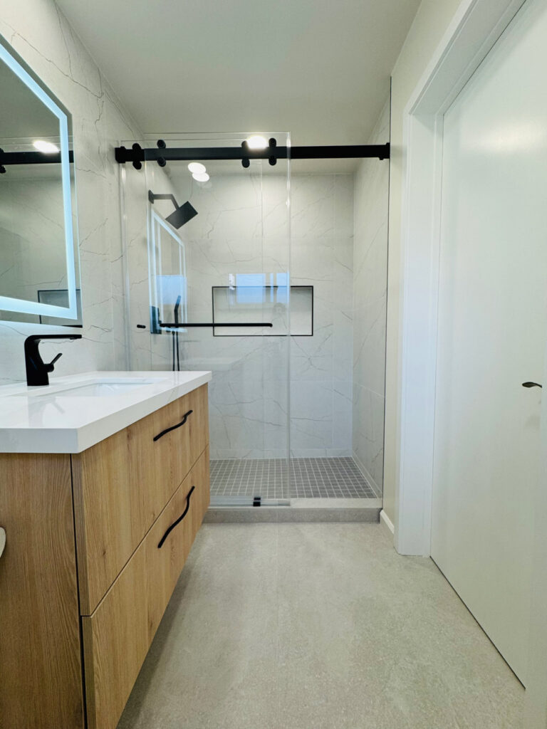 San Jose bathroom remodel master bath viewed toward the shower, with a light oak floating vanity and black wavy pulls, a backlit mirror, and a glass shower on a black rail with gray square mosaic flooring over concrete-look tile.