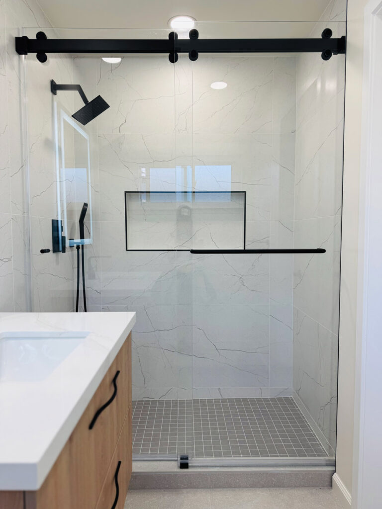 San Jose bathroom remodel shower with a black roller rail, matte black rain head and handheld, a shampoo niche trimmed in black Schluter profile and a towel bar, and a gray square mosaic base set within porcelain walls with soft veins.