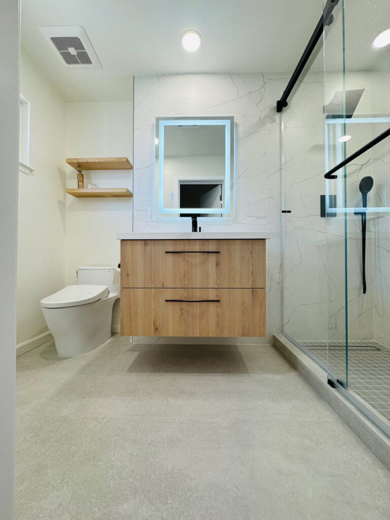 San Jose bathroom remodel with an oak floating vanity and lit mirror, twin oak floating shelves, a bidet toilet, and the glass shower with black fittings at right.