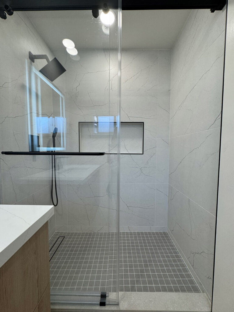 San Jose bathroom remodel shower viewed through the glass on its black roller rail, with a black rain head, a shampoo niche trimmed in black Schluter profile and towel bar, and a gray square mosaic base draining to a linear channel.
