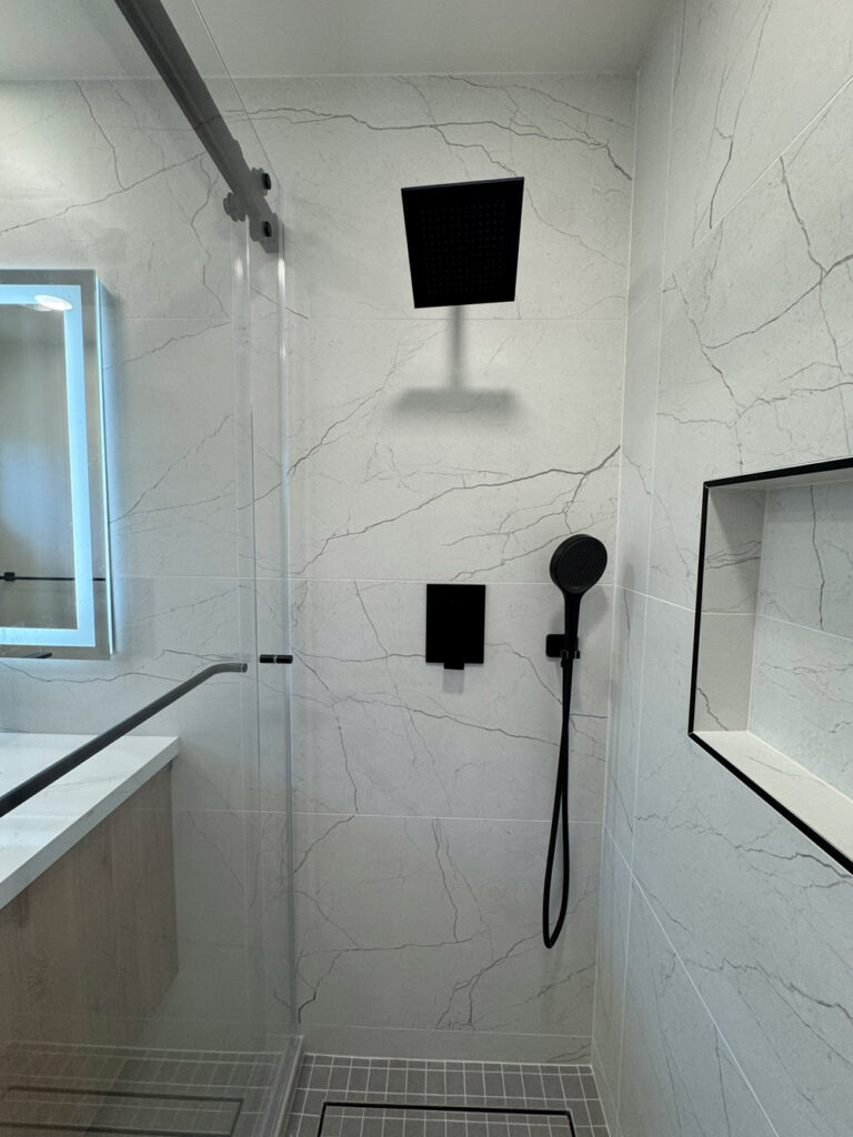 San Jose bathroom remodel shower fittings with a large matte black square rain head, a flush black valve plate and a round handheld on a black hose, set against porcelain with dramatic veins beside the shampoo niche.